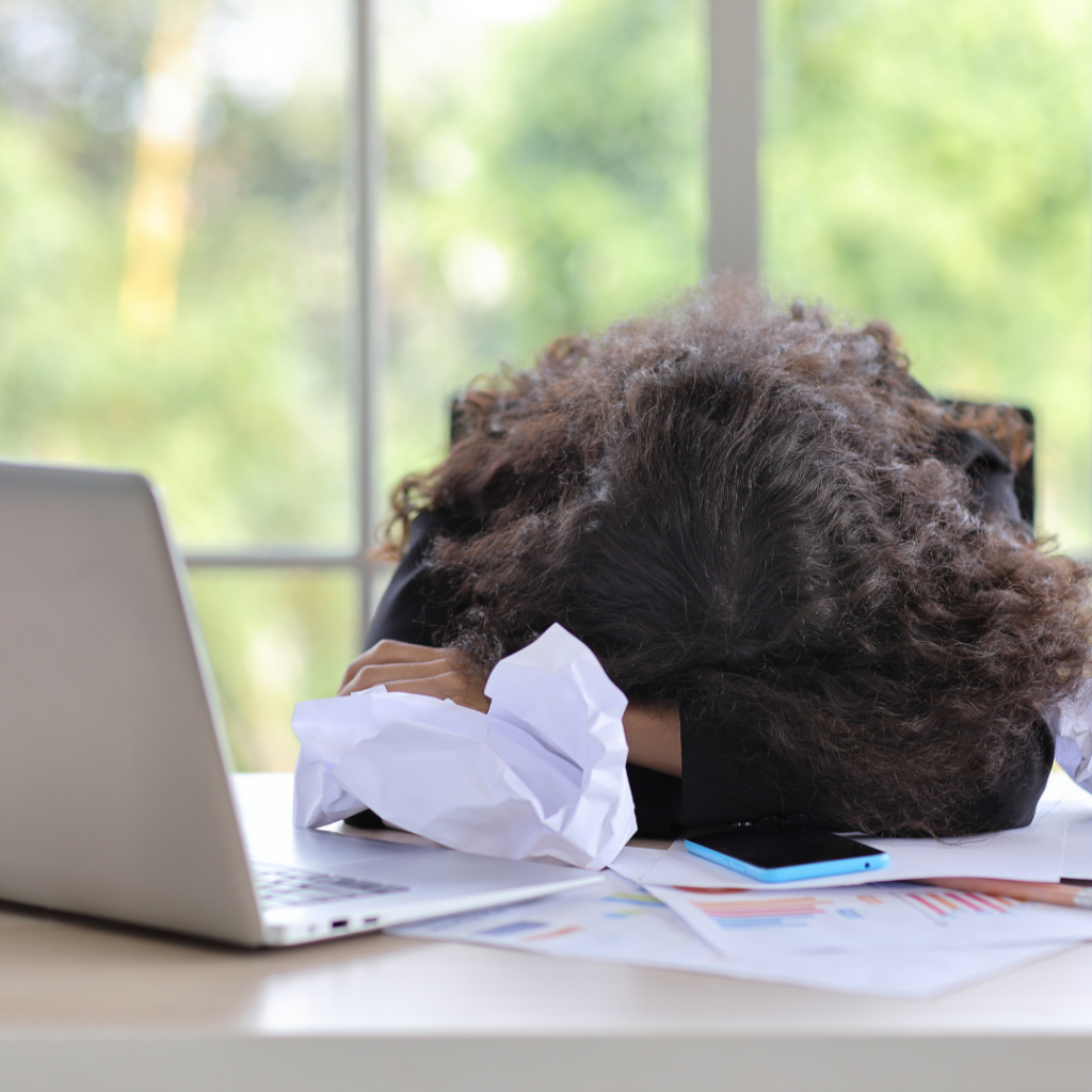 A person with curly hair, wearing a black blazer, is slumped over their desk with their head resting on their arms in exhaustion. Crumpled papers, a laptop, a smartphone, and scattered documents with colorful charts are spread across the desk. A large window in the background lets in natural light, showing green foliage outside.