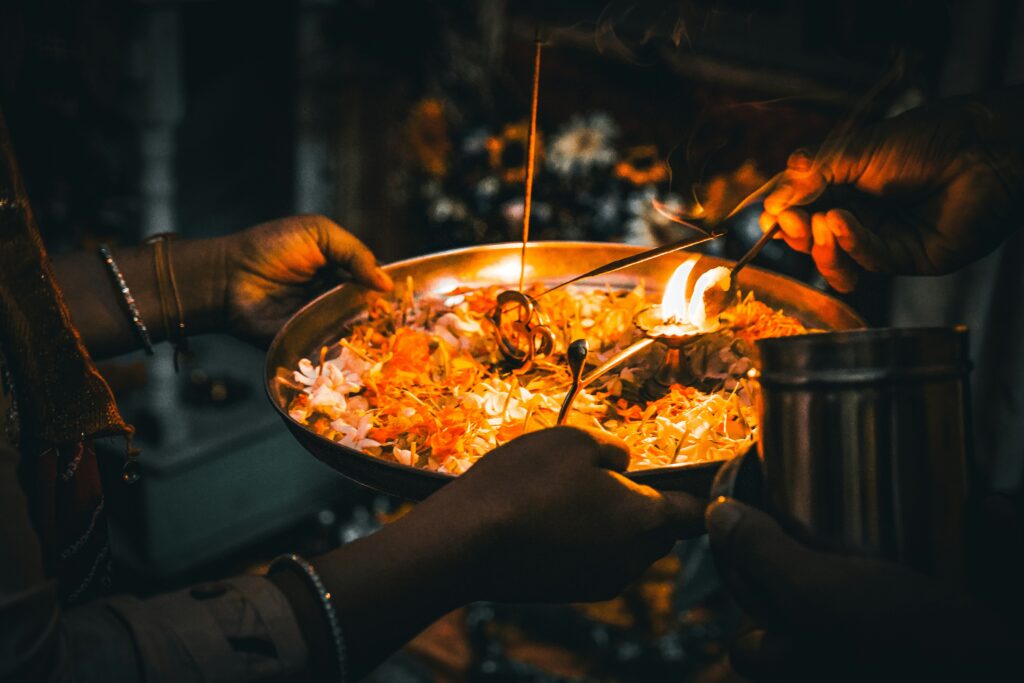 The image shows a close-up view of hands performing Aarti, a Hindu ritual of worship. A circular metal plate, likely brass or copper, holds offerings such as flower petals and what appears to be grains or herbs. A small flame, possibly fueled by ghee or camphor, burns brightly in the center of the plate, radiating a warm glow.